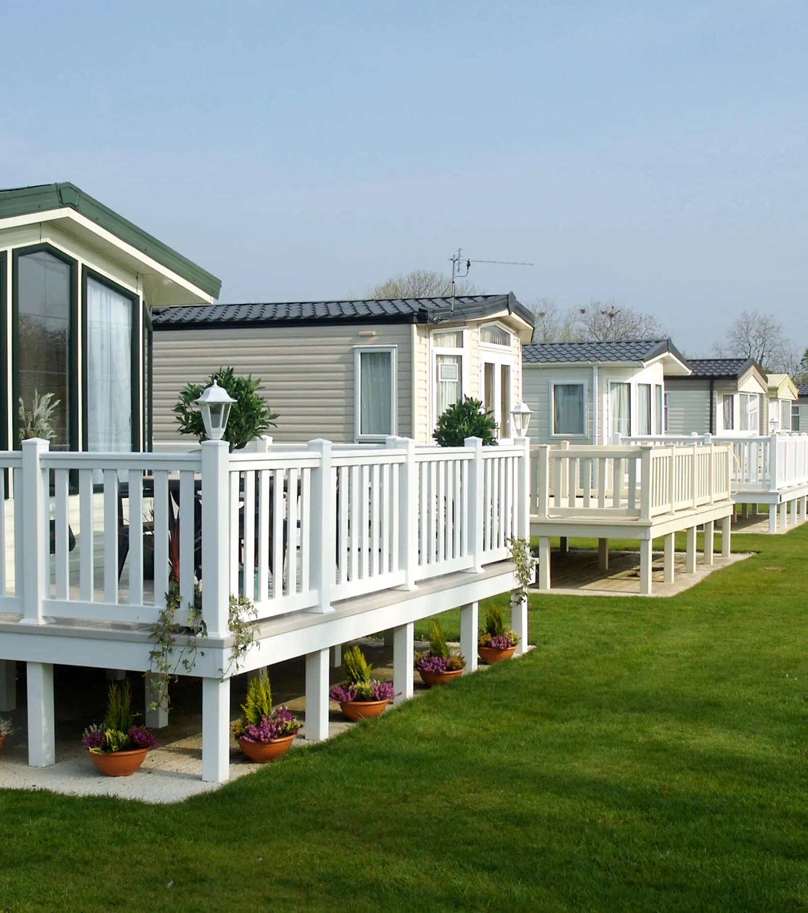 A row of mobile homes, showcasing pristine white wooden decks and railings adorned with potted plants, boasts premium mobile home parts. Nestled on a well-maintained lawn beneath a clear sky, these residences exemplify comfort and style.