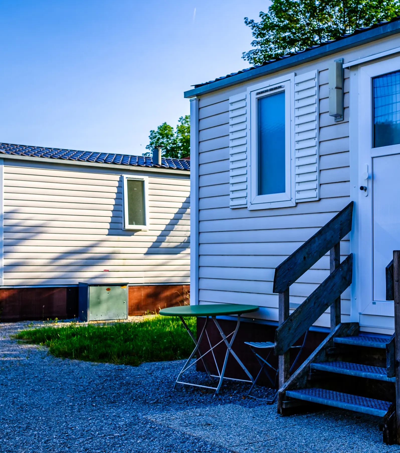 Two mobile home rentals with white siding are nestled in a grassy area. A green table and steps grace the foreground, inviting relaxation.