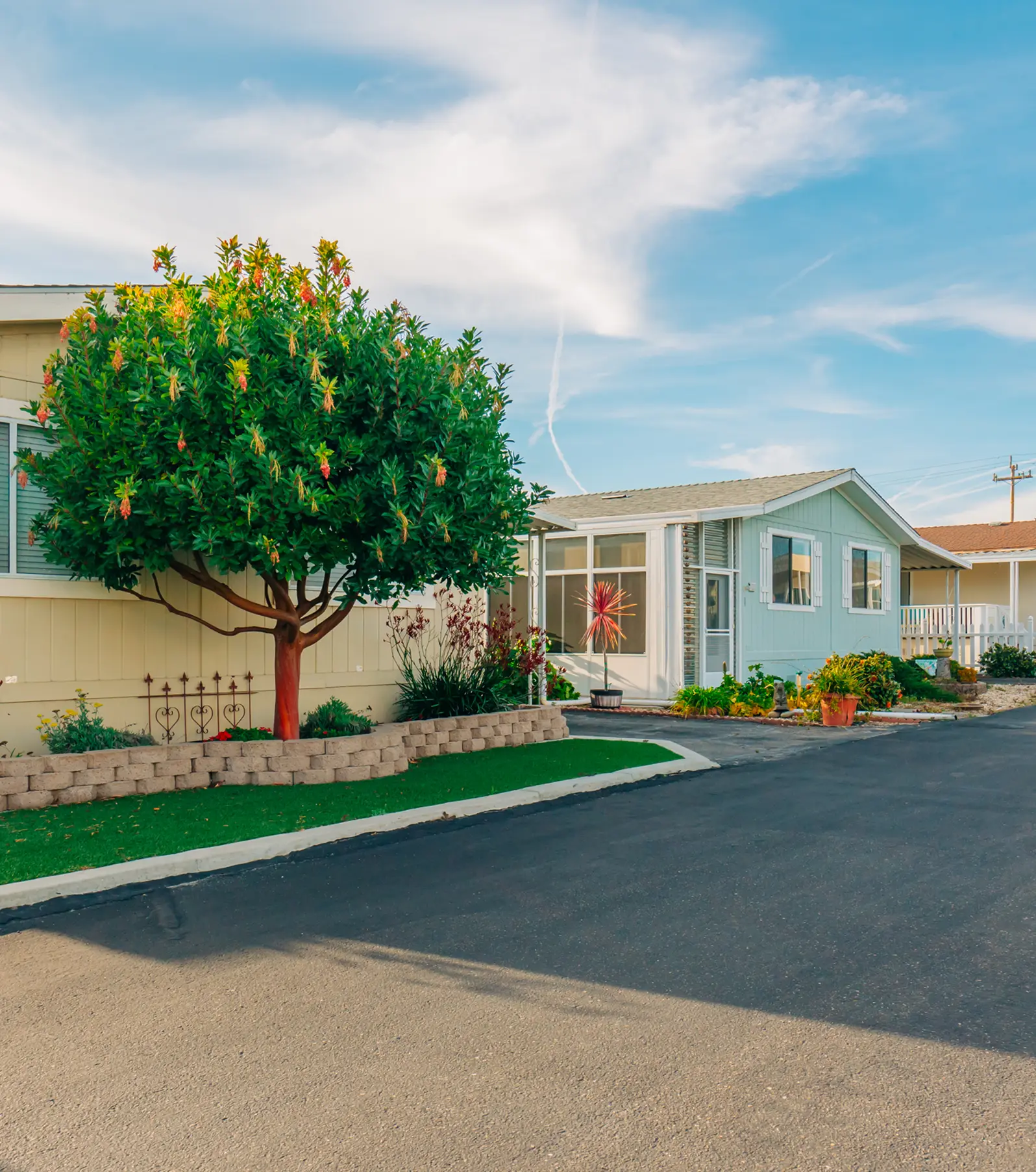 A small, light blue mobile home with large windows and a paved driveway is bordered by a green lawn and a colorful flowering tree under a clear blue sky.